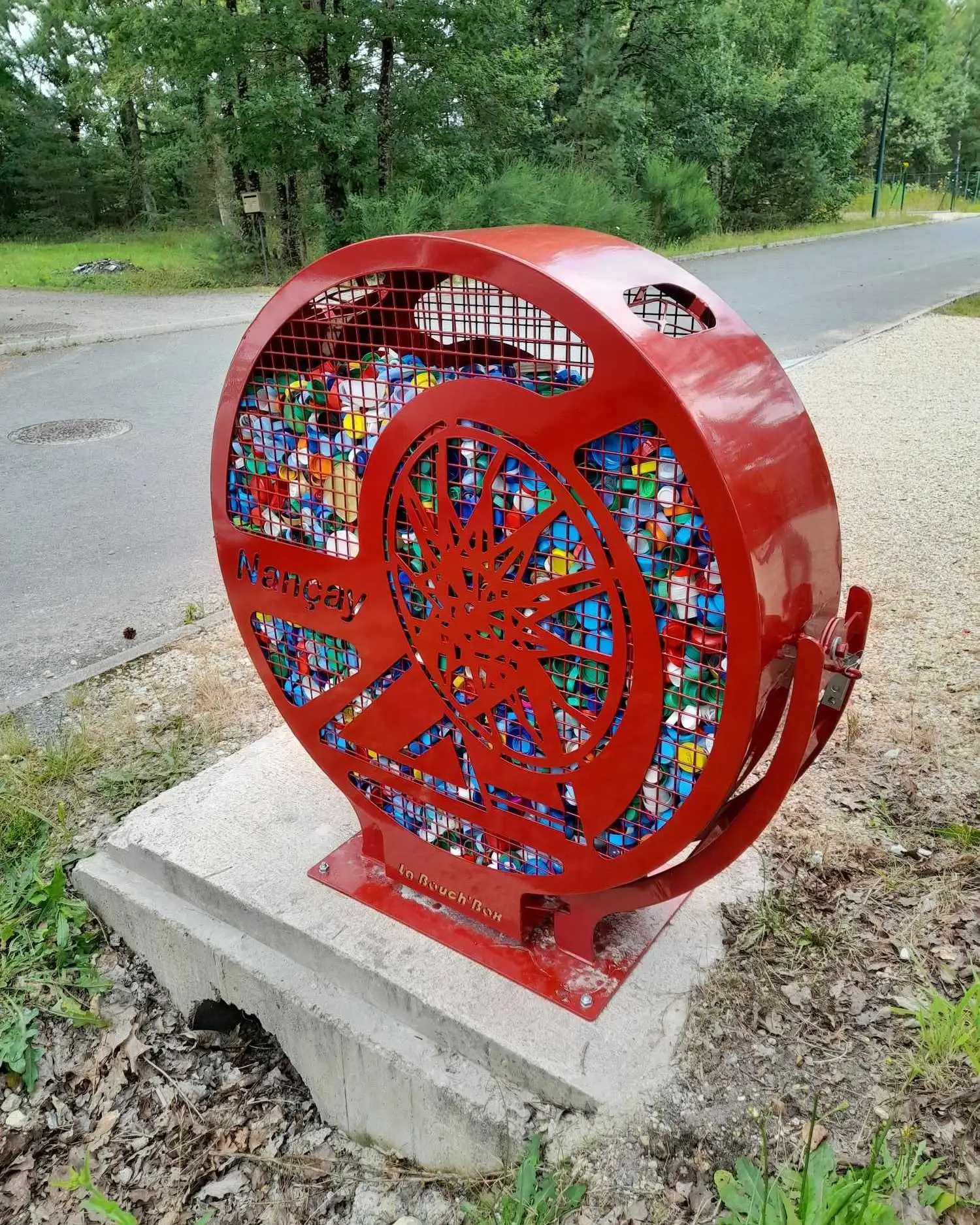 Bouch’Box rouge personnalisée au nom de Nançay, remplie de bouchons plastique colorés, posée sur un massif béton